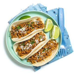 A teal plate with three soft chicken tacos for family dinner with limes on a white countertop.