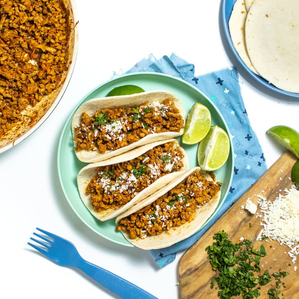 A spread for Family dinner with a teal plate with three soft tacos with ground, chicken and limes in the middle, a skillet of ground, chicken, meat, a plate of tortillas, and a cutting board with cilantro cheese and lime.
