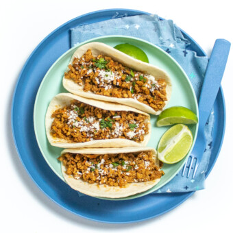 Two blue kids plates with three soft tacos with ground chicken, cheese, and cilantro, with limes on the plates on a white countertop.