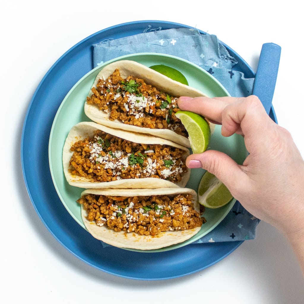 Two blue plates with three soft tacos with ground, chicken and spices with cheese and cilantro, with a hand, squirting line onto the tacos on the white countertop.