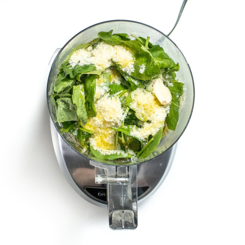 A food processor on the white countertop with spinach basil, Parmesan, lemon juice, and avocado, getting ready to be mixed together.