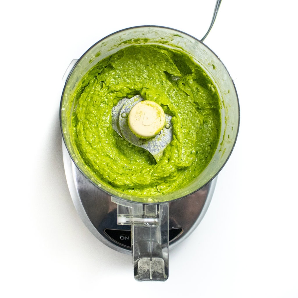 A silver food processor on a white countertop with a avocado, spinach pesto sauce mixed together.