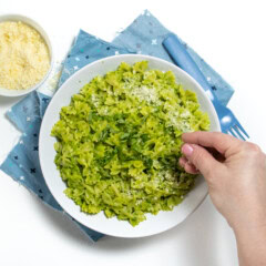 White bowl with bowtie pasta with a creamy avocado, spinach pesto sauce with a hand putting Parmesan on top. The bowl is sitting on a blue napkin on a white countertop with a small bowl of Parmesan next to it.
