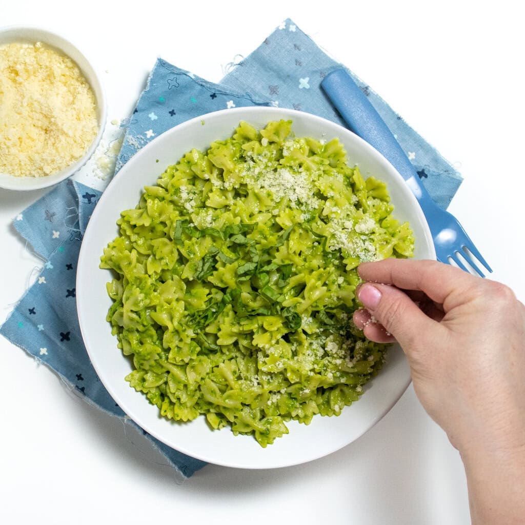 White bowl with bowtie pasta with a creamy avocado, spinach pesto sauce with a hand putting Parmesan on top. The bowl is sitting on a blue napkin on a white countertop with a small bowl of Parmesan next to it.