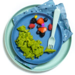 Blue kids plates with green scrambled eggs, freshly cut fruit and a blue fork and blue napkin on a white countertop.