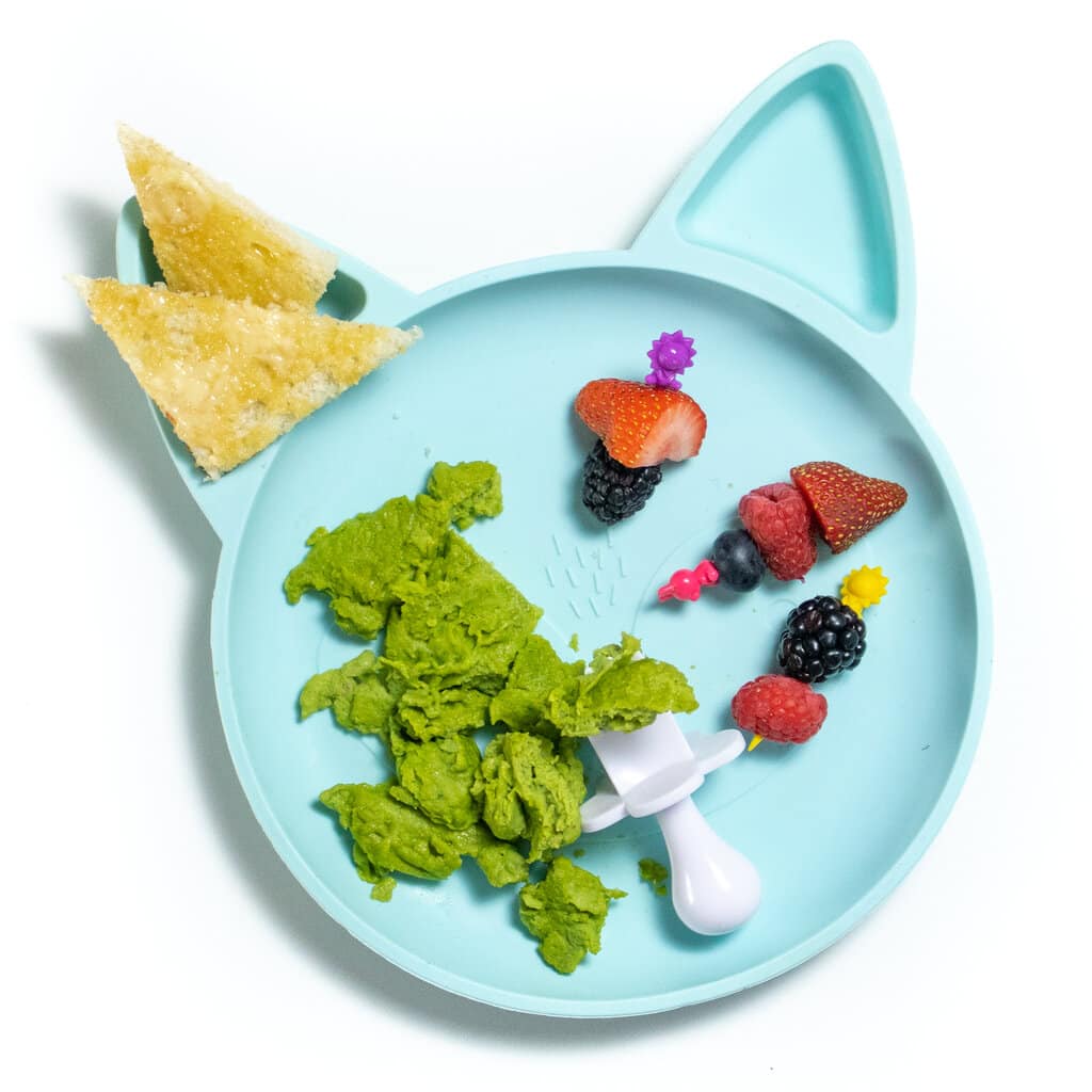 A blue toddler play that has sections with green, scrambled eggs, freshly cut fruit and two triangles of toast on a white countertop.