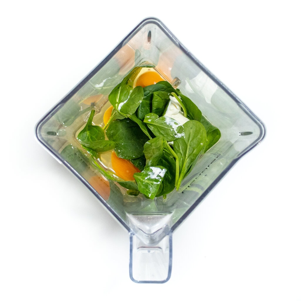 A clear blender, full of ingredients, like spinach, eggs, cream cheese, on a white countertop.