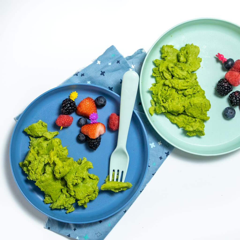 Two blue kids plates with green scrambled eggs, a blue fork and blue napkin and a white cutter top and freshly cut fruit.