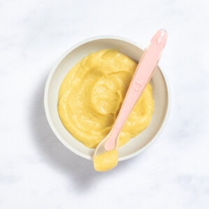 Gray baby bowl sitting on a white marble counter with parsnip puree inside, with a pink baby spoon resting on the rim with puree inside.