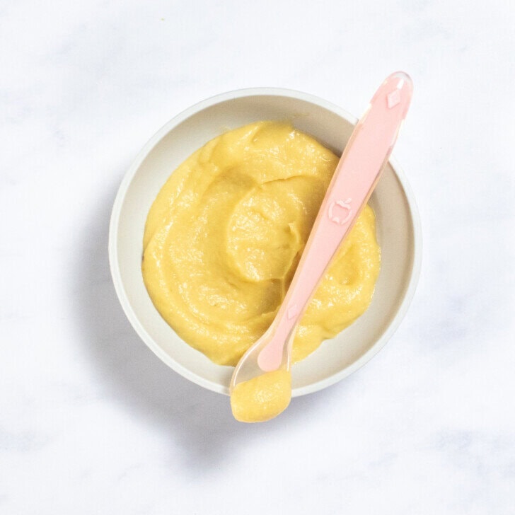 Gray baby bowl sitting on a white marble counter with parsnip puree inside, with a pink baby spoon resting on the rim with puree inside.