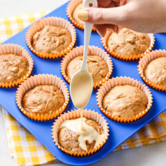 Hand holding a white spoon over a blue muffin tin full of carrot cake muffins. The hand is drizzeling cream cheese icing over the front muffin.