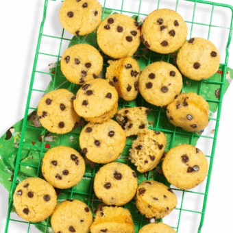 Green cooling rack with a bunch of mini chocolate chip muffins resting on top. There is a green kid themed napkin underneath to catch the crumbs and they are sitting on a marble kitchen counter.