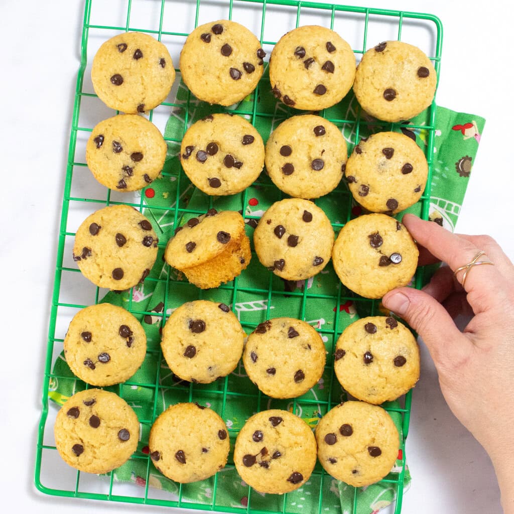 A green wire cooling rack on a green napkin with a kid print sitting on my white marble kitchen counter. On the cooling rack is mini chocolate chip muffins with my hand reaching towards one. 
