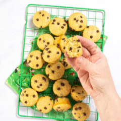 My hand holding up a half eaten mini chocolate chip muffin over a green cooling rack full of mini muffins. The rack is resting on a green kid-themed napkin on my white kitchen counter.