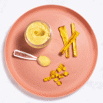 Pink baby plate on a white marble counter with different ways to serve parsnips to your baby - puree, strips for baby-led weaning and chopped for finger foods.