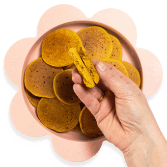 My hands holding a baby cereal pancakes torn into two halves over a pink baby plate full of small pancakes.