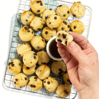 My hand holding a mini pancake bite over a small bowl of maple syrup that is resting on a cooling rack along with dozens of other mini pancake bites.