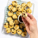 My hand holding a mini pancake bite over a small bowl of maple syrup that is resting on a cooling rack along with dozens of other mini pancake bites.