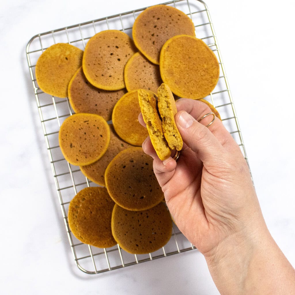 My hand holding a baby pancakes over a cooling rack filled with golden brown pancakes for baby-led weaning. 