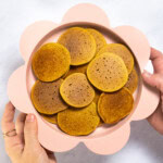 My hands holding a pink baby plate full of small baby cereal pancakes over my white marble kitchen counter.