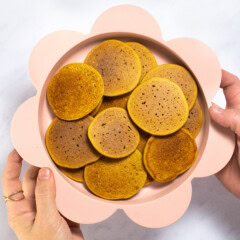 My hands holding a pink baby plate full of small baby cereal pancakes over my white marble kitchen counter.