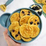 My hand holding a kids blue bear plate full of avocado pancakes. Some of them have blueberries in them. The plate is over my white marble kitchen counter with a cooling rack with more pancakes on them.