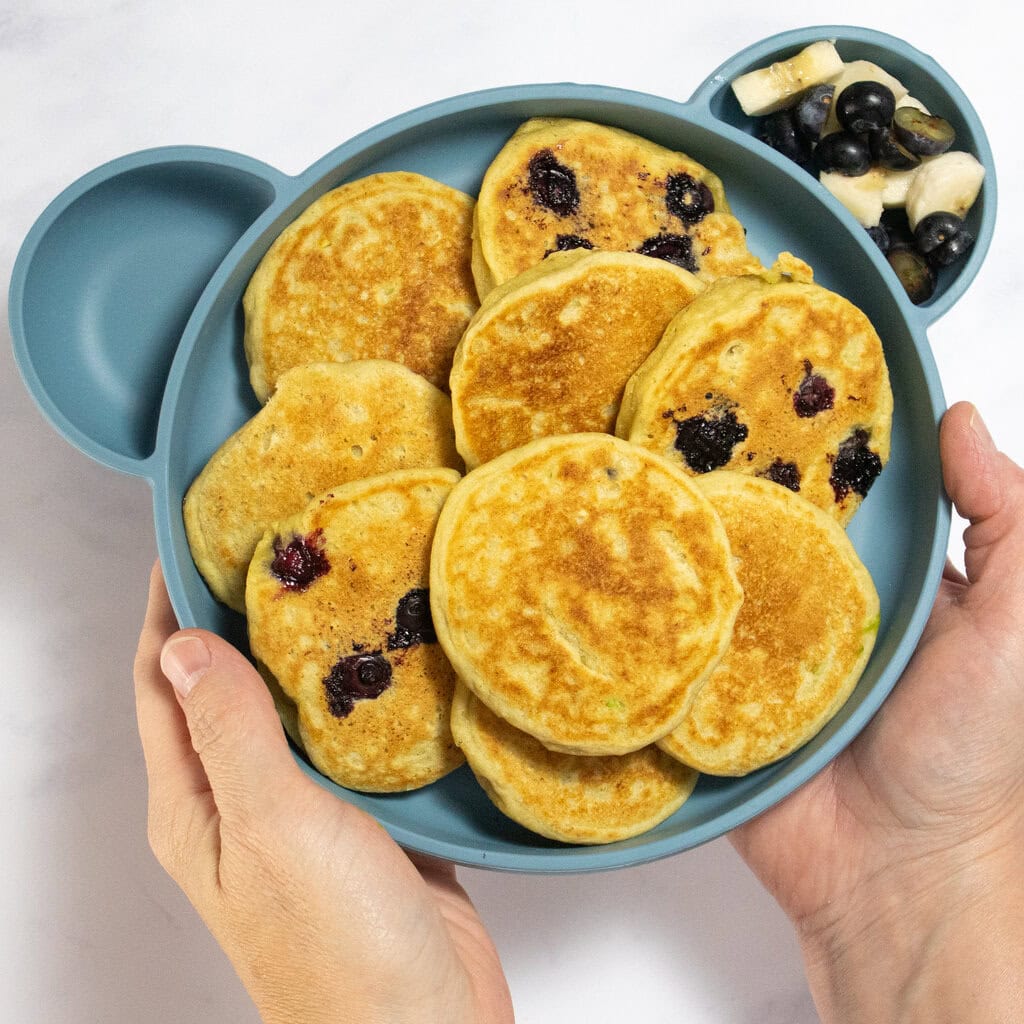 My hands holding a blue kids plate full of avocado banana pancakes. Some of the pancakes have blueberries. The plate is over my white marble kitchen counter with a cooling rack full of pancakes as well. 