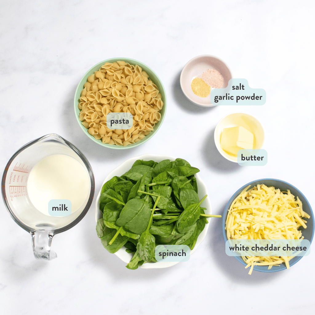 Ingredients for green mac and cheese on my white marble kitchen counter in small bowls - milk, spinach, shredded cheddar cheese, butter, pasta and spices.