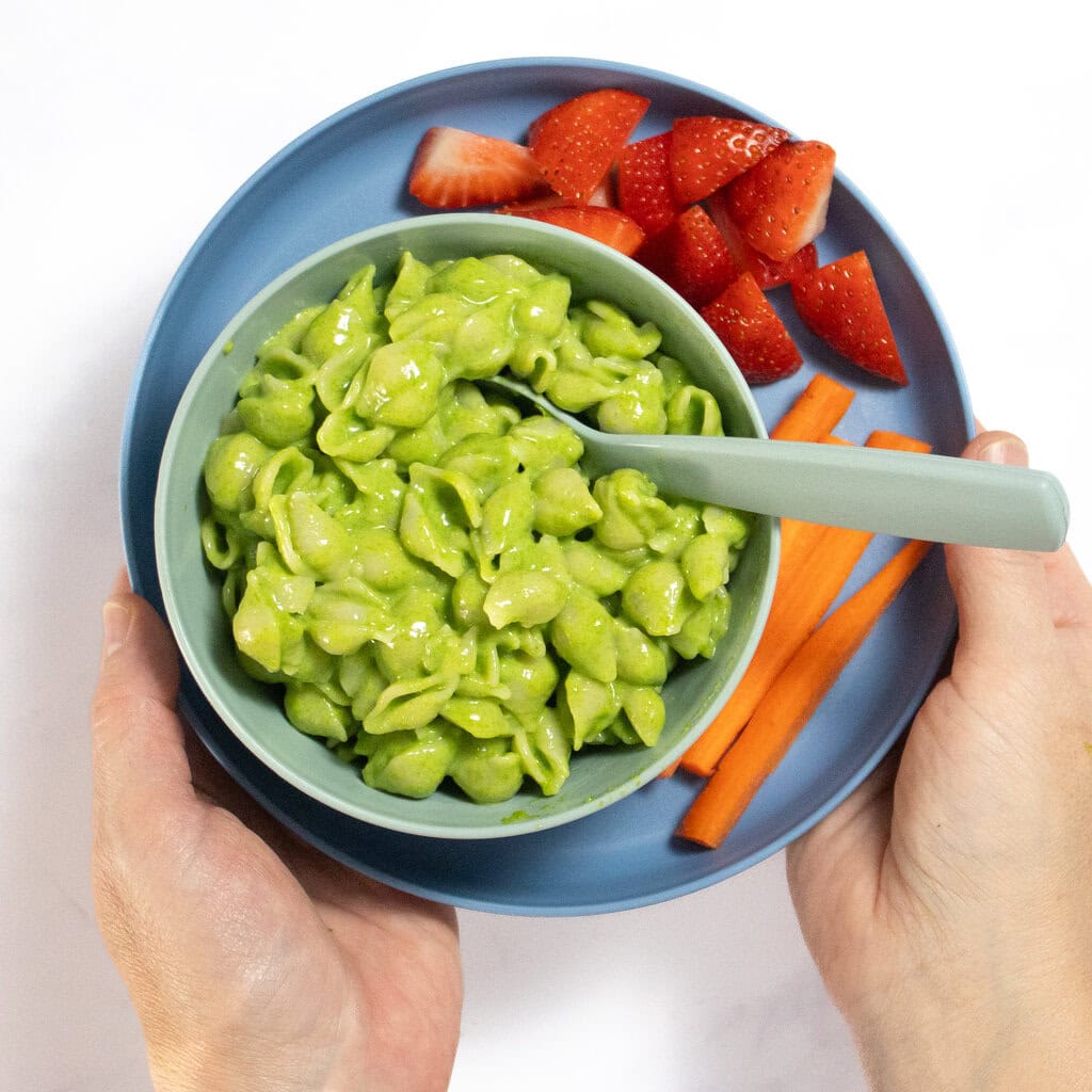 my two hands holding a blue kids plate with green mac and cheese, cut strawberries and slices of carrots.