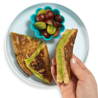 My hands holding a spinach grilled cheese slice over a blue kids plate with a small bowl of cut green and purple grapes.