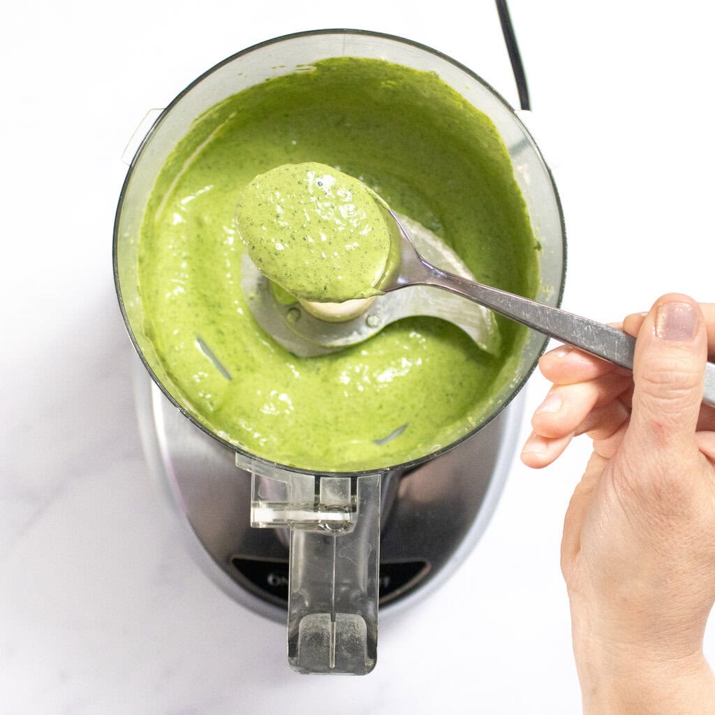 A food processor on my kitchen counter with my hand holding a spoonful of spinach mixture for the grilled cheese.
