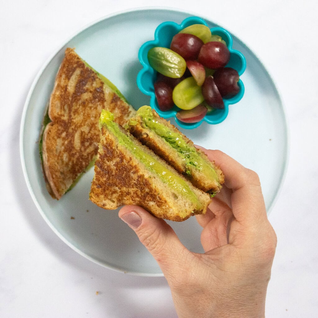 My hand holding two quarters of grilled cheese with spinach over a blue plate.
