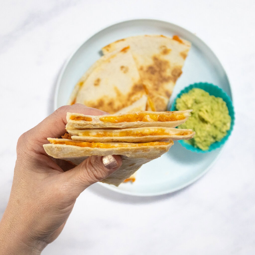 My hand holding three wedges of sweet potato quesadillas over a blue kids plate on my kitchen counter.