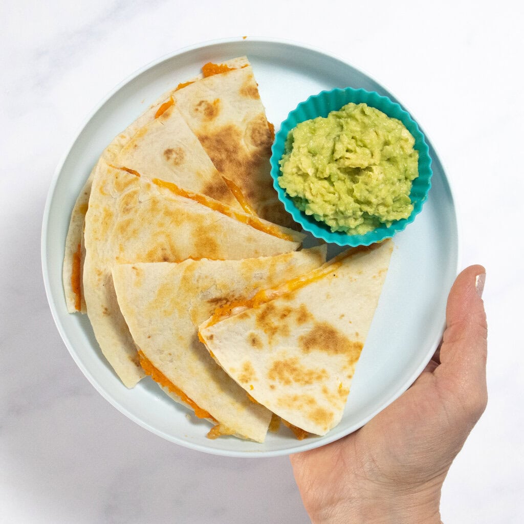 My hand holding a blue plate with wedges of sweet potato quesadillas with a side of guacamole in a small kids bowl.