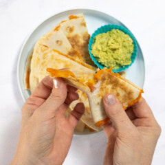 My hands pulling a sweet potato quesadilla apart over a blue kids plate with more wedges of quesadilla.