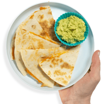 My hand holding a blue plate with wedges of sweet potato quesadillas with a side of guacamole in a small kids bowl.