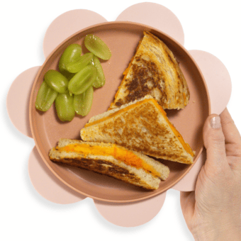 My hand holding a pink kids plate over my kitchen counter that has wedges of grilled cheese and grapes cut lengthwise.