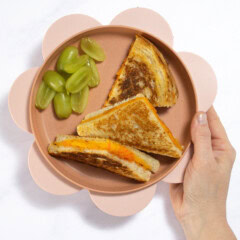 My hand holding a pink kids plate over my kitchen counter that has wedges of grilled cheese and grapes cut lengthwise.