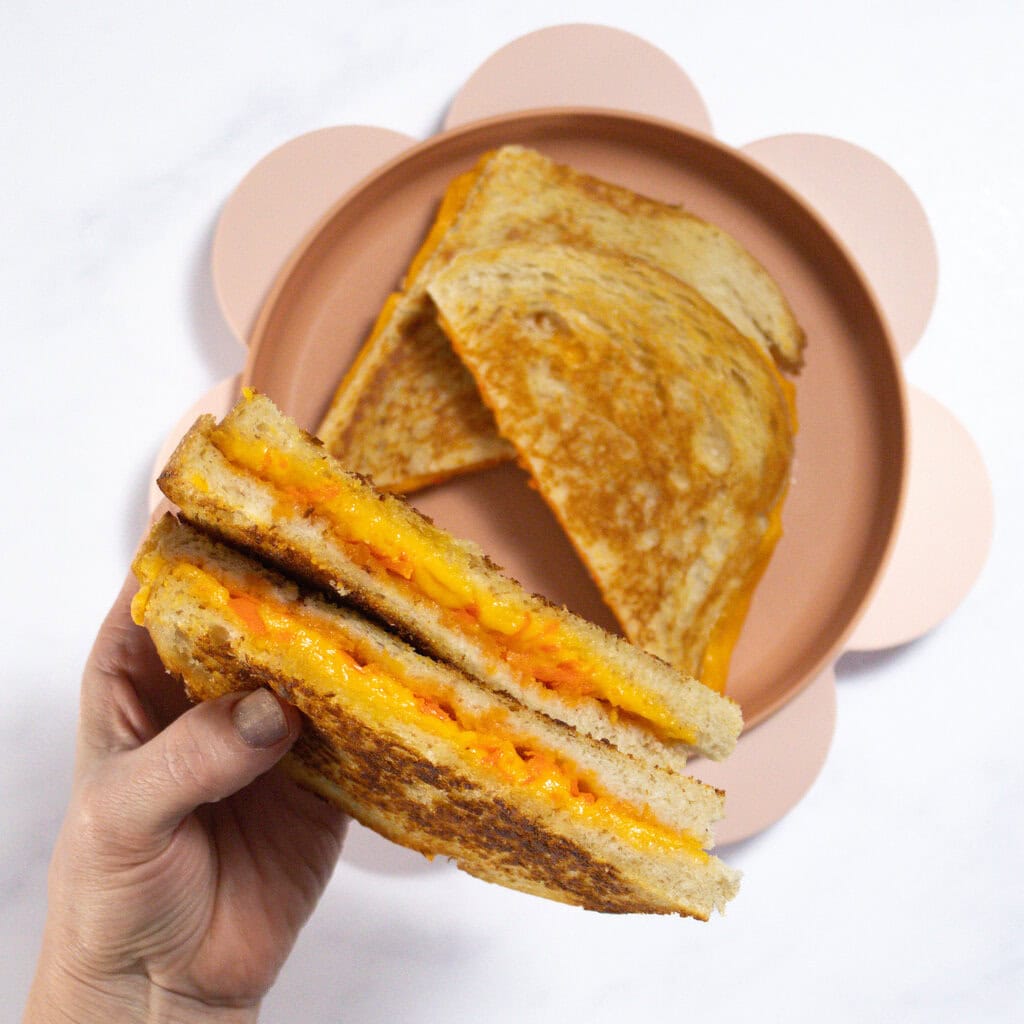 My hands holding wedges of grilled cheese over a pink plate of grilled cheeses.
