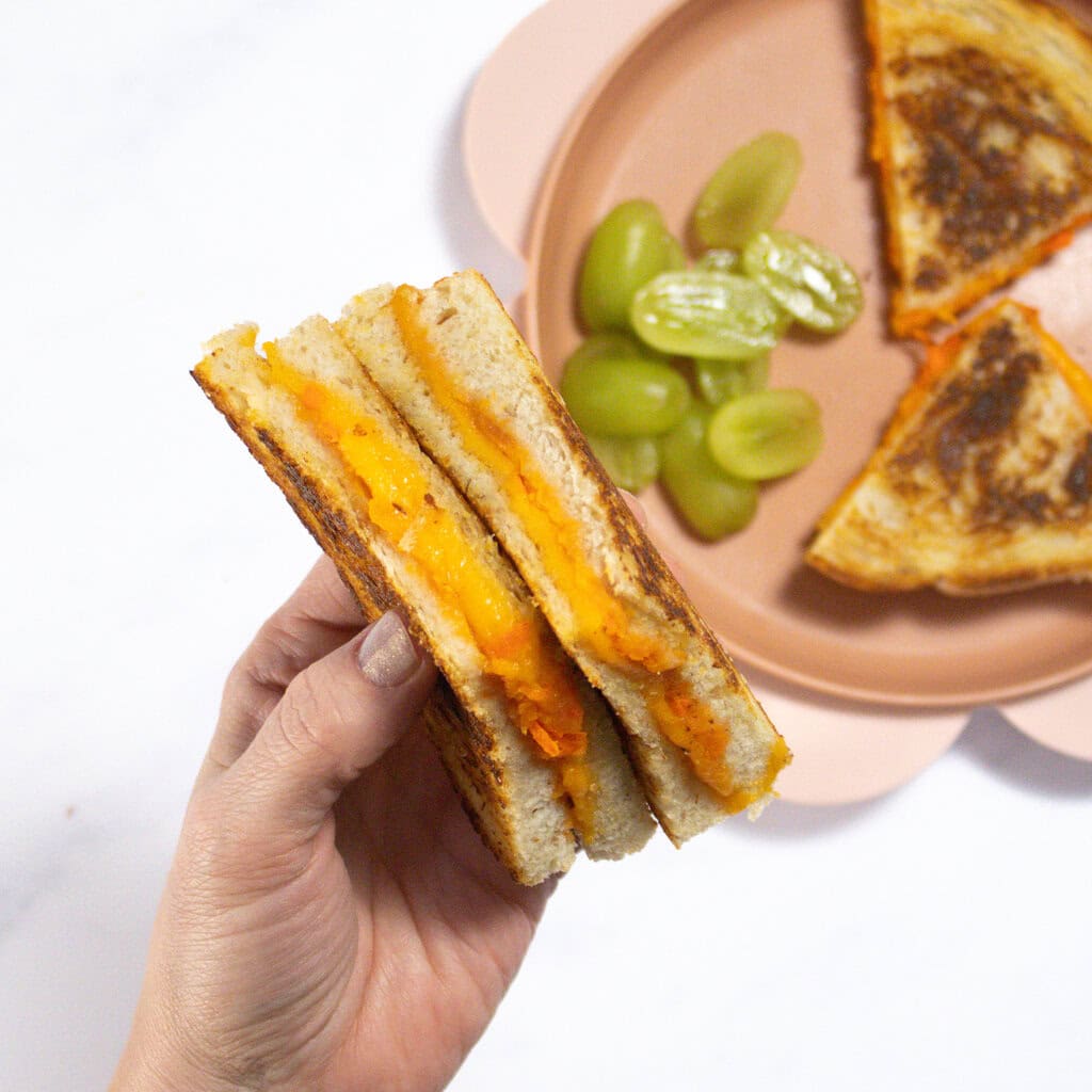My hand holding 2 wedges of veggie grilled cheese over a pink kids plate with more grilled cheese and grapes.