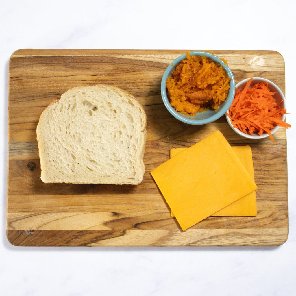 A wood cutting board sitting on my white kitchen counter with the ingredients for veggie grilled cheese - bread, slices of cheese, mashed sweet potato and shredded carrots.