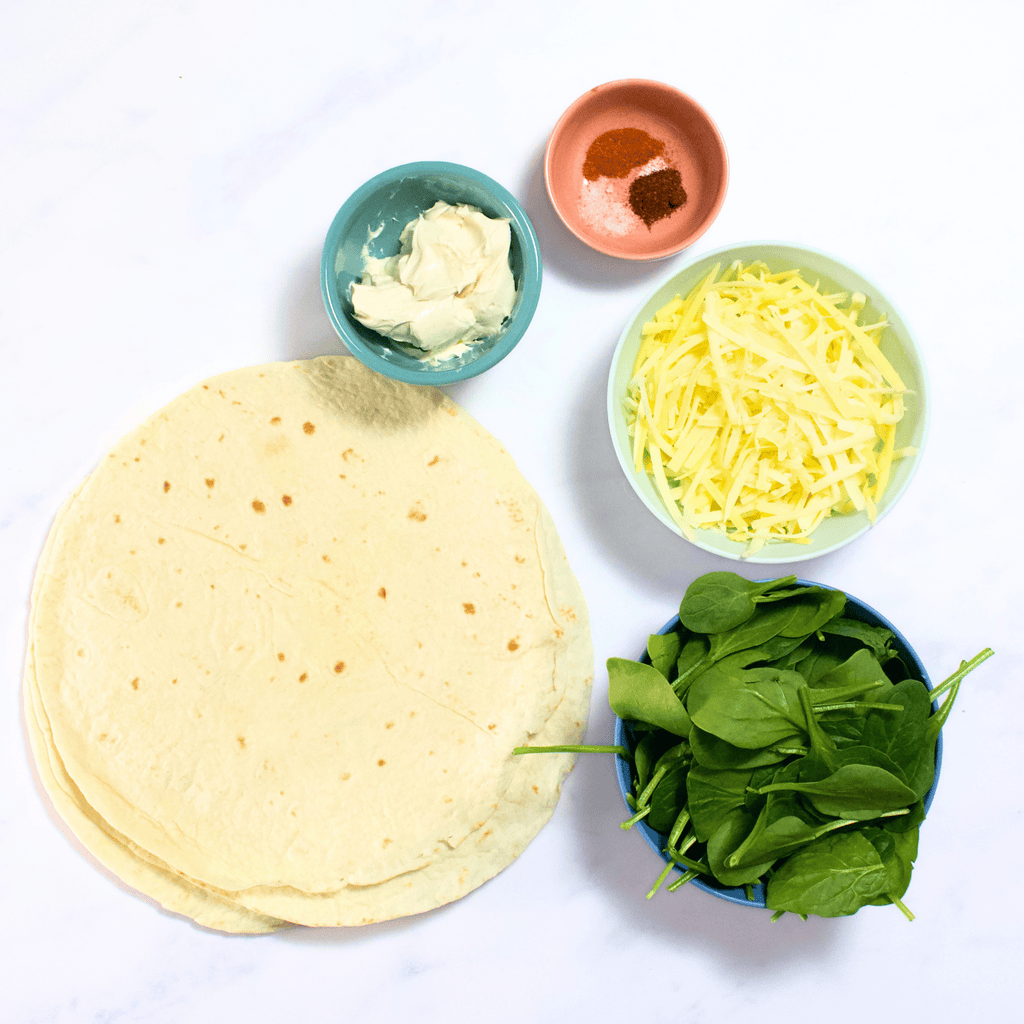 Ingredients for the spinach green quesadillas on my white kitchen counter - tortillas, spinach, white cheddar cheese, cream cheese, and a small bowl of spices.