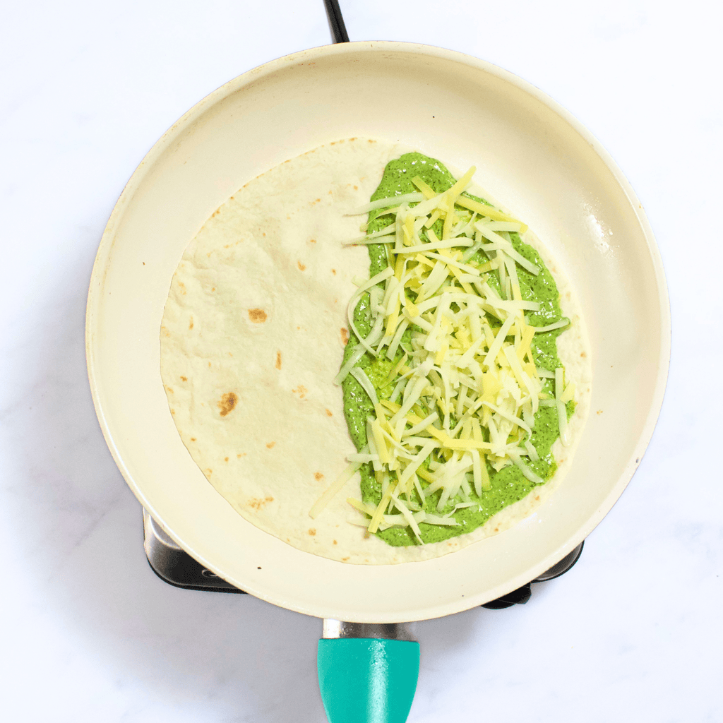 White skillet with a tortilla, green spinach spread and white cheddar getting ready to be flipped.