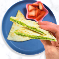 My hand holding two wedges of a spinach quesadilla over a blue plate with more wedges and a small container of cut strawberries all sitting on my white countertop.